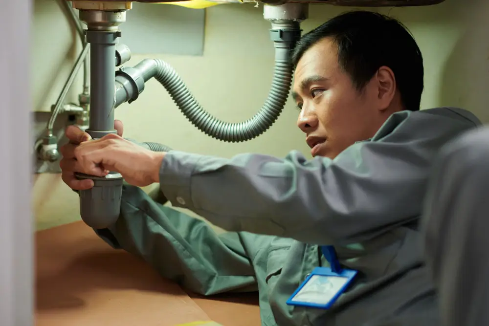 A plumber passes a tool to another under a kitchen cabinet, demonstrating the labor that contributes to why emergency plumber kenmore are expensive but worth every penny