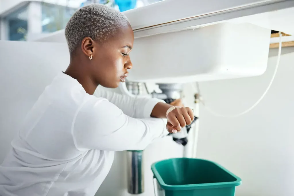 Woman fixing a blocked drain Ashgrove under a kitchen sink with a green bucket placed below for water collection