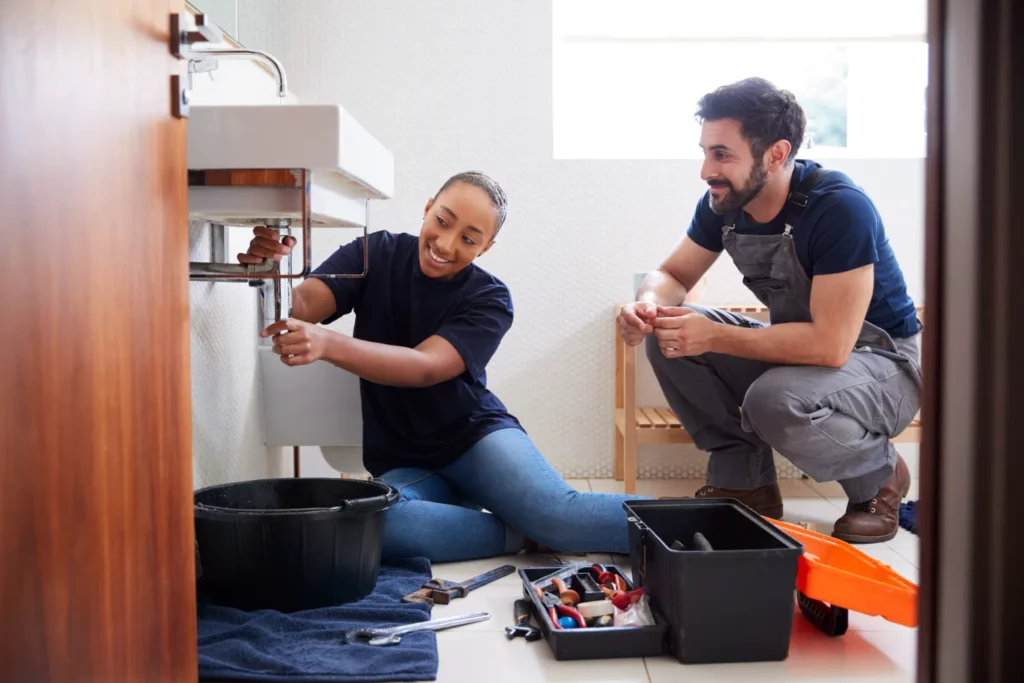 An experienced plumber teaches an apprentice under a sink, showing the expertise that factors into why plumbers are so expensive