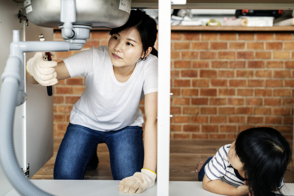 Focused woman inspecting under-sink plumbing, demonstrating how often should drains be inspected, with a child observing the process.
