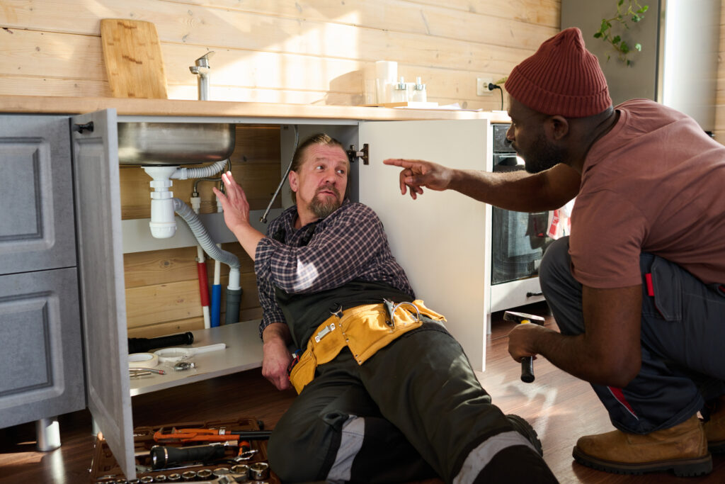 A plumber lies under a kitchen sink working on pipes while another man kneels nearby, pointing and advising. Tools are scattered around them, emphasizing the need for a skilled local emergency plumber for quick repairs.