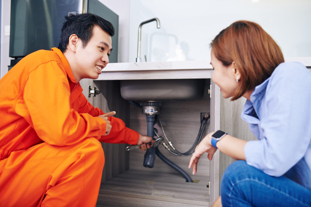 A smiling plumber in an orange jumpsuit from an emergency plumbing company fixes a pipe under a kitchen sink while a woman kneels beside him, attentively watching the repair process.